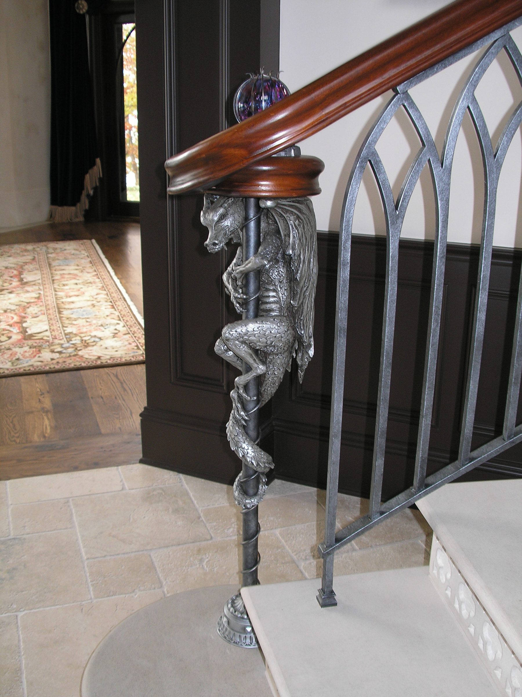 A cast-metal dragon coiled around the base of a French Gothic staircase newel post, finished to match the wrought-iron pickets and topped by a glass orb.