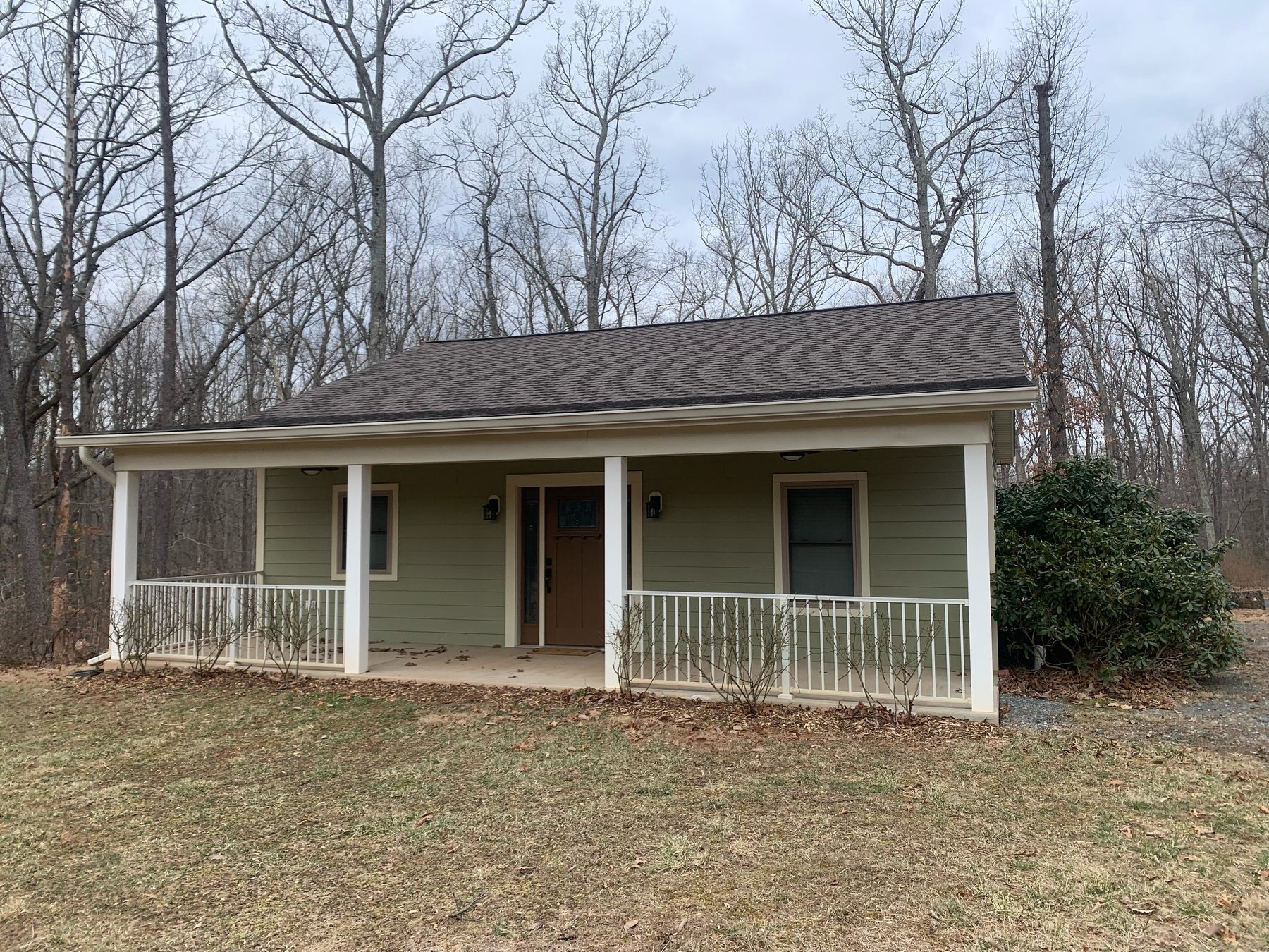 The single-storey guest house — green lap siding, white-railed covered porch and a gabled roof, set in bare winter woods.